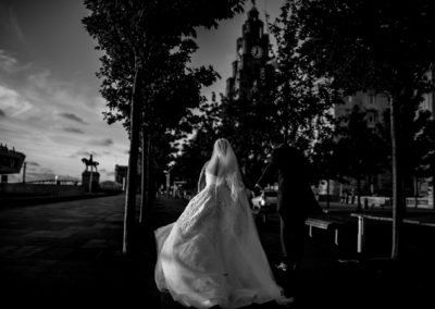 Black and white documentary photograph of bride and groom walking towards the Liver Buildings in Liverpool
